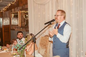 Speaker addresses guests at formal event venue with decorative lighting and white curtain backdrop.
