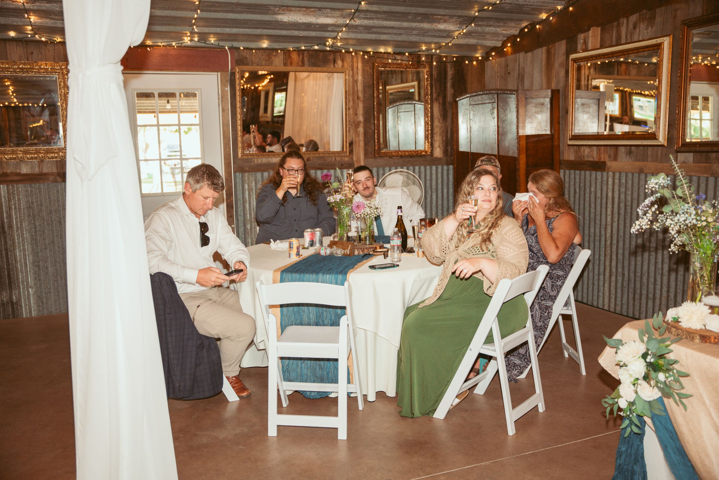 A rustic wedding reception inside a barn venue with people seated at decorated tables with string lights overhead.