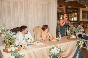 Wedding reception table decorated with floral centerpieces and white drapery in rustic indoor venue.