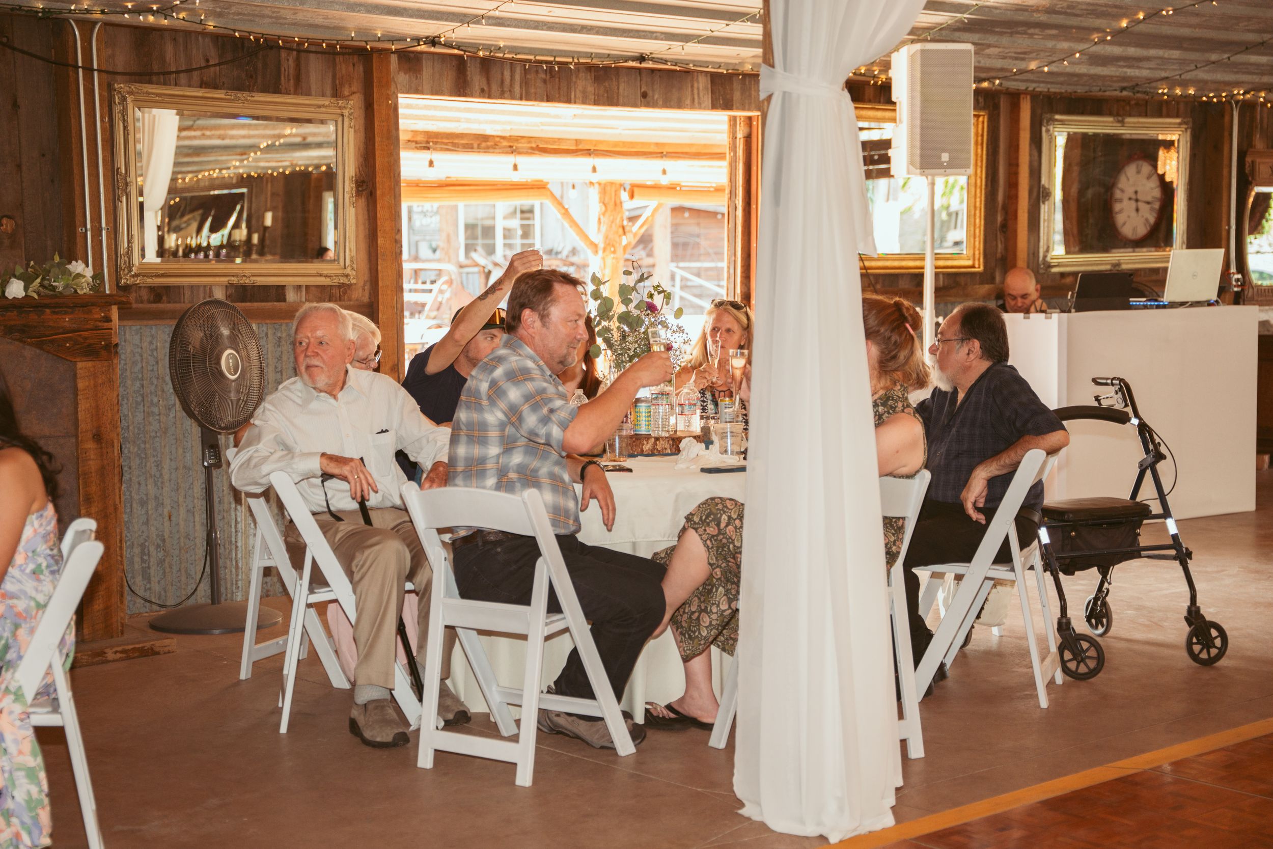 Wedding reception guests converse at white folding chairs inside a rustic wood-paneled venue.