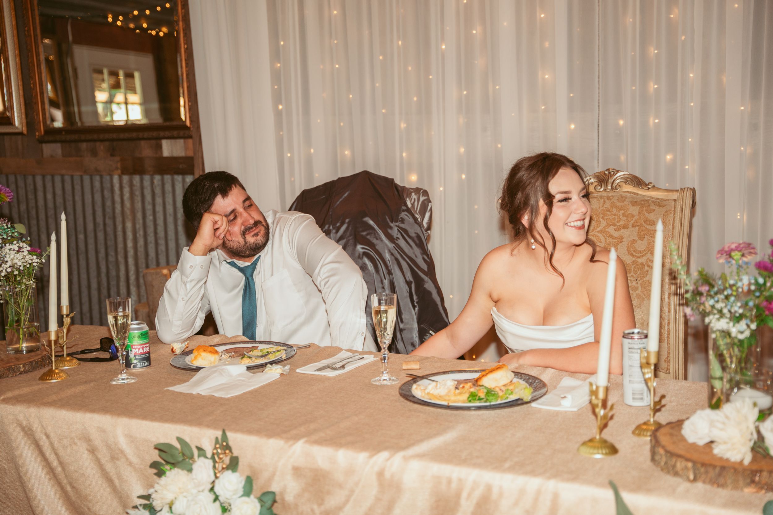 Wedding reception guests enjoy a candlelit dinner at rustic wooden tables with elegant white decor and twinkle lights above.