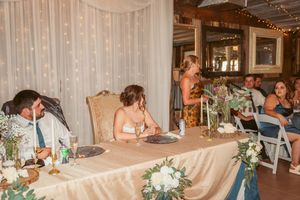 Wedding reception scene at a rustic venue with white floral centerpieces and string lights overhead during dinner service.