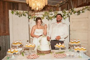 Wedding dessert table with rustic decor features tiered white cake, cupcakes and treats under crystal chandelier.