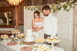 People cutting into a multi-tiered white wedding cake with pastries and desserts displayed at rustic reception venue.