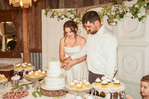 Couple cutting their wedding cake at dessert table adorned with cupcakes and greenery.