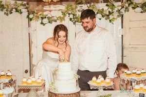 Wedding couple cuts into their elegant white tiered cake while surrounded by floral decorations at reception venue.