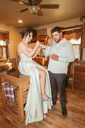 Couple in formal attire share a sweet moment while sipping drinks together in a rustic interior with ceiling fan.