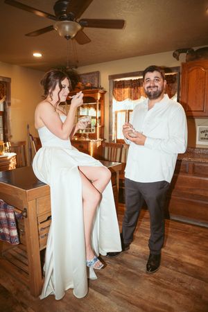 Two people in formal attire share a moment in a rustic kitchen space with wooden furniture and ceiling fan.
