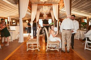 Wedding reception inside a rustic venue with wooden floors, white drapes, and a crystal chandelier hanging overhead.