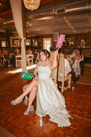 A bride in a white satin gown plays beer pong at a wedding reception on a wooden dance floor under string lights.