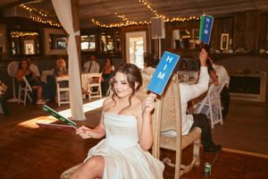 A person in a white wedding dress holds up blue paddle signs during a reception event with string lights overhead.