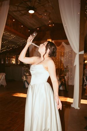 A person in a white wedding dress takes a drink from a bottle during a reception with string lights and white drapes.