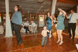 Wedding guests dance together on a rustic wooden dance floor under string lights and white drapes at a reception venue.