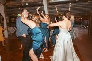 Wedding reception guests and the bride dance together on the dance floor at an indoor venue with twinkle lights.