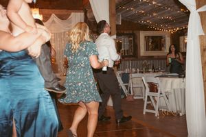 Wedding guests dance together on a wooden floor decorated with string lights and white fabric drapes at a reception venue.