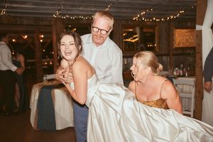 Wedding party dances joyfully under string lights in a barn-style venue with elegant chandeliers.
