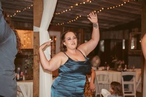A person in a teal satin dress dances joyfully at an indoor reception venue with string lights.