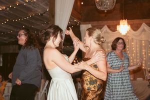 Wedding guests dance together on a rustic wood floor under string lights and chandeliers at a reception venue.
