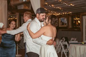 Wedding guests dance and embrace on a rustic indoor dance floor decorated with twinkling string lights overhead.