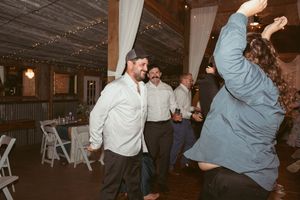 People dancing energetically at a casual reception in a barn-style venue with string lights overhead.