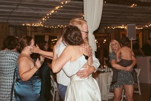 Joyful dancing and hugging at a wedding reception under twinkling lights in a rustic venue.