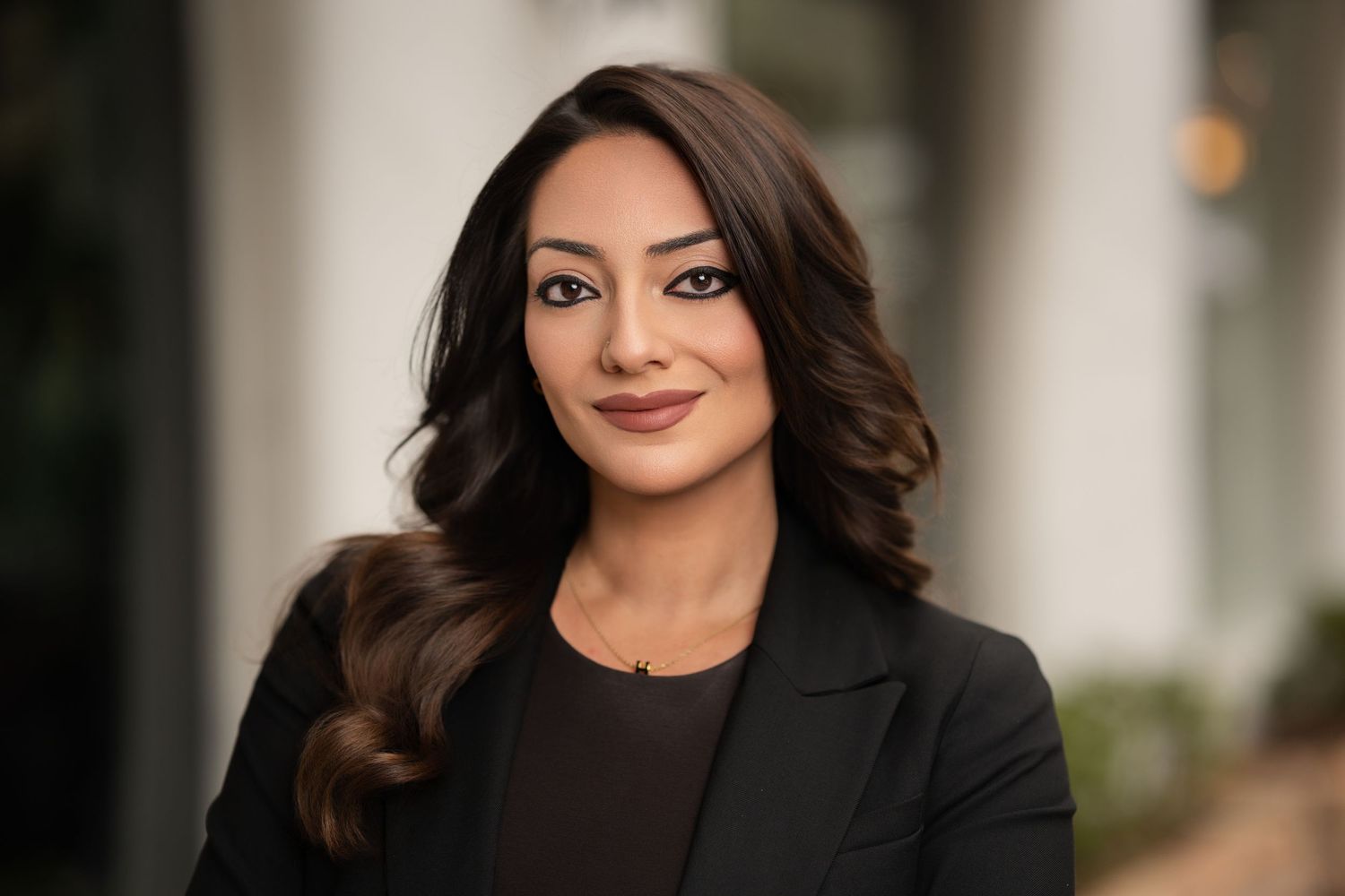 Professional woman in black blazer poses outdoors with soft bokeh background for corporate headshot.