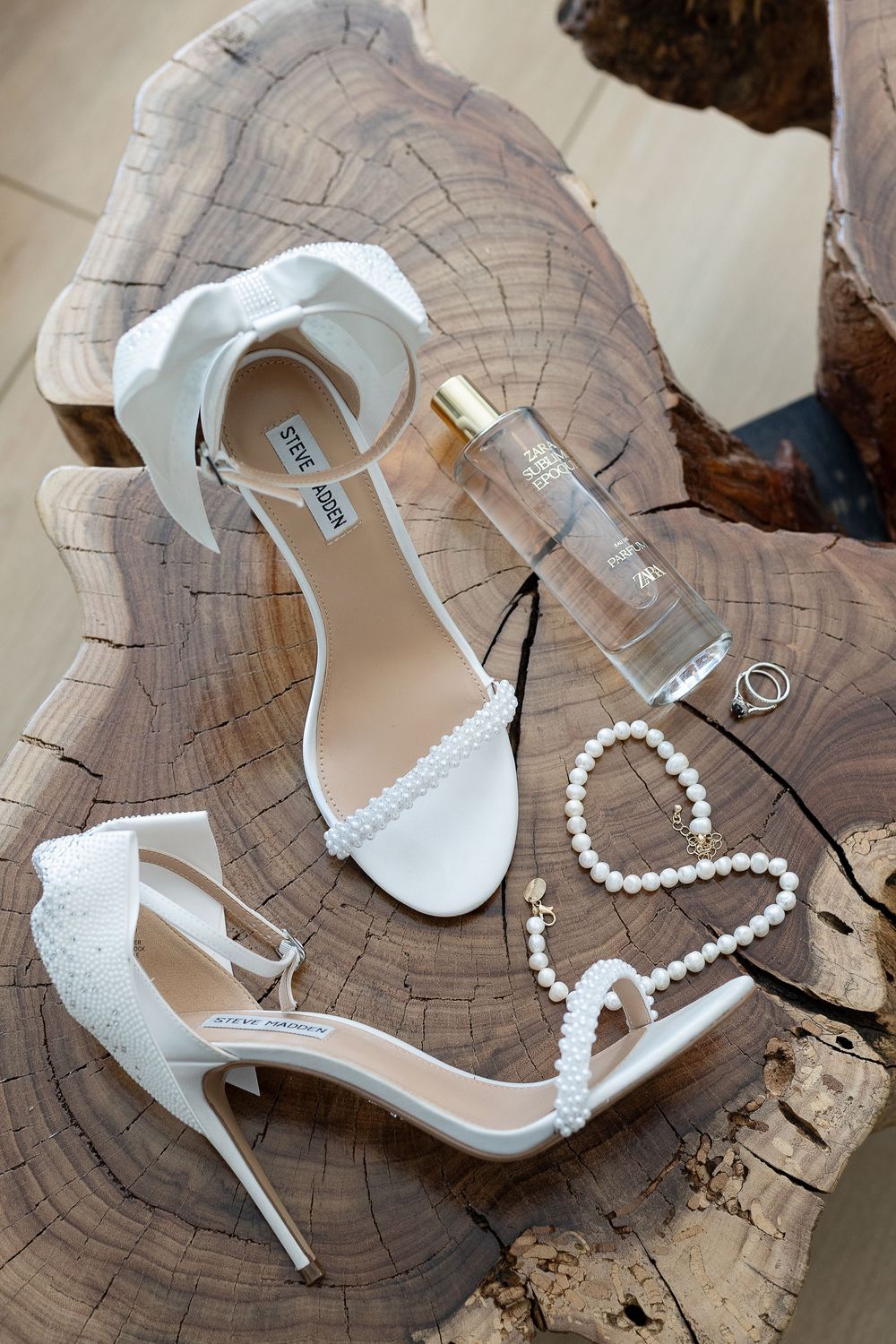 White wedding accessories including Steve Madden heels, pearl necklace and perfume displayed on rustic wood surface.