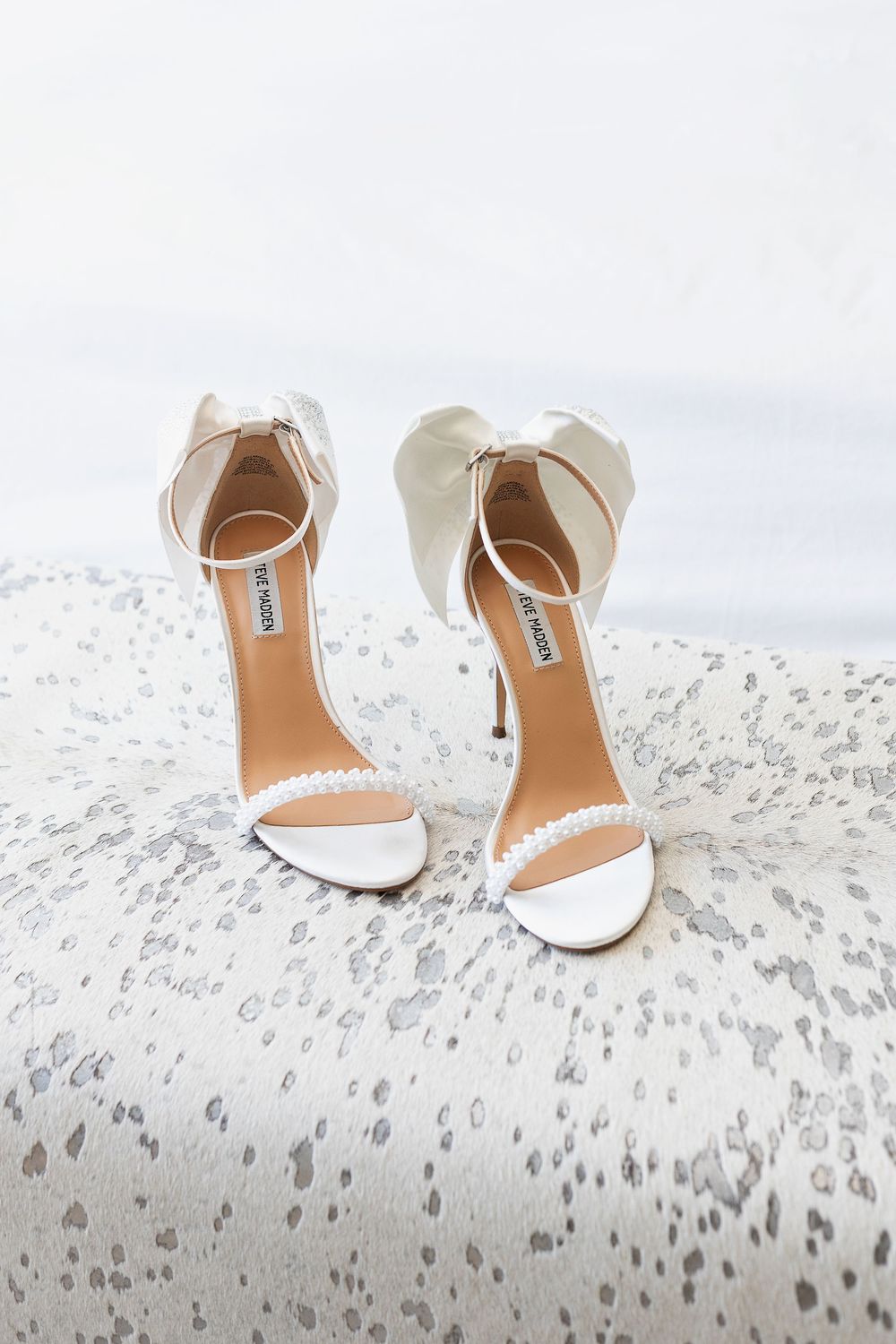 White bridal high heel sandals with ankle straps displayed on a marbled surface.