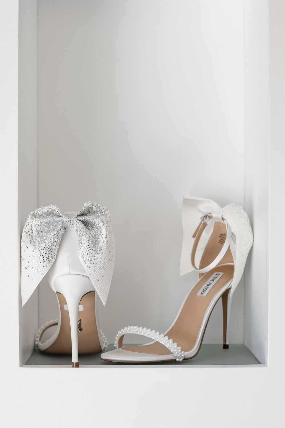 Elegant white bridal heels with crystal embellishments and bows displayed in a minimalist white shelf setting.