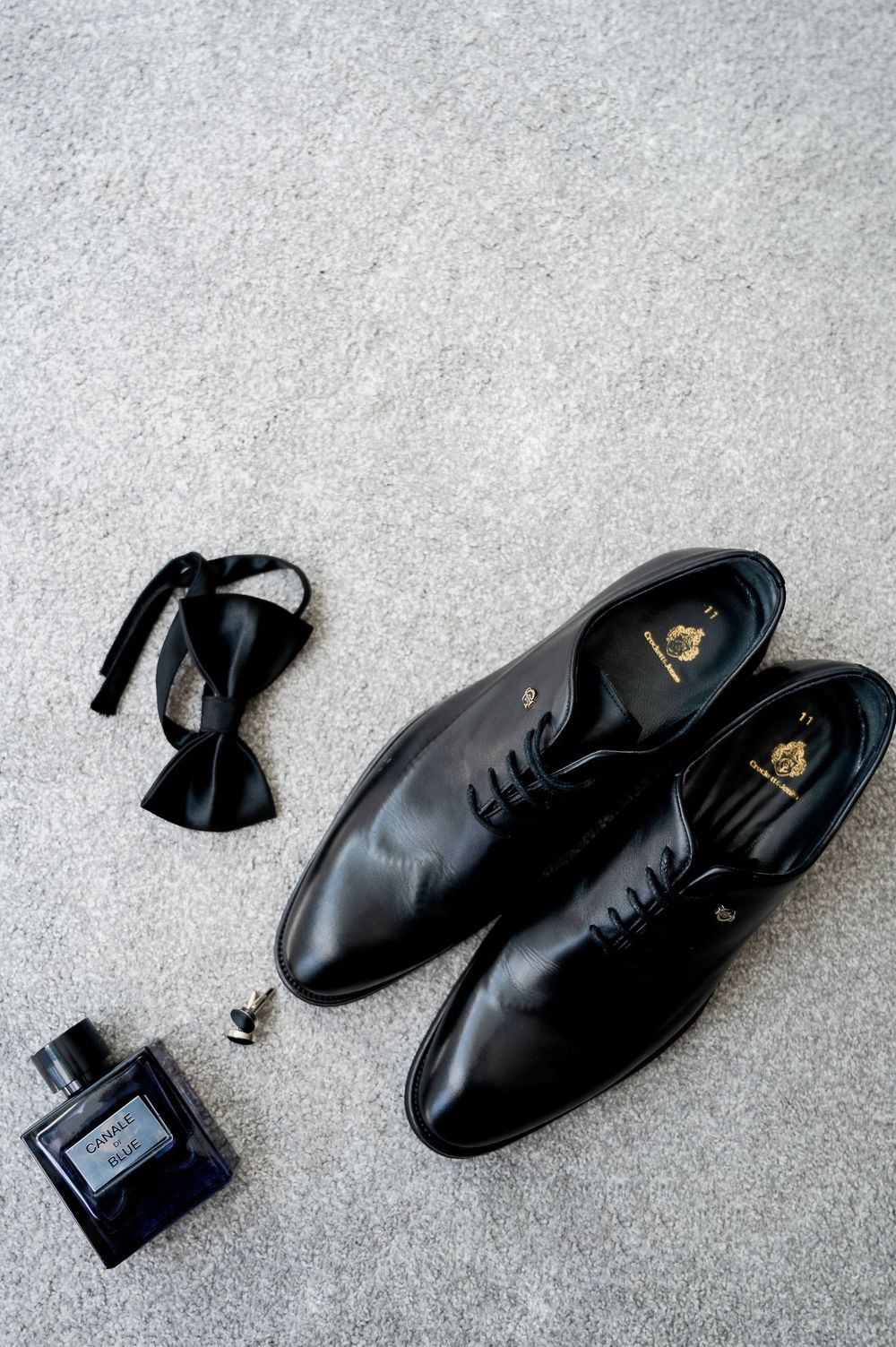 Black formal men's dress shoes with bow tie and perfume bottle arranged on gray carpet.