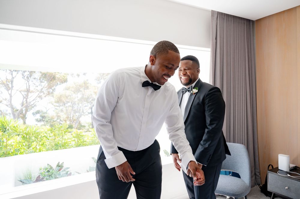 Two people in formal black and white attire share a laugh while getting ready in a bright hotel room.