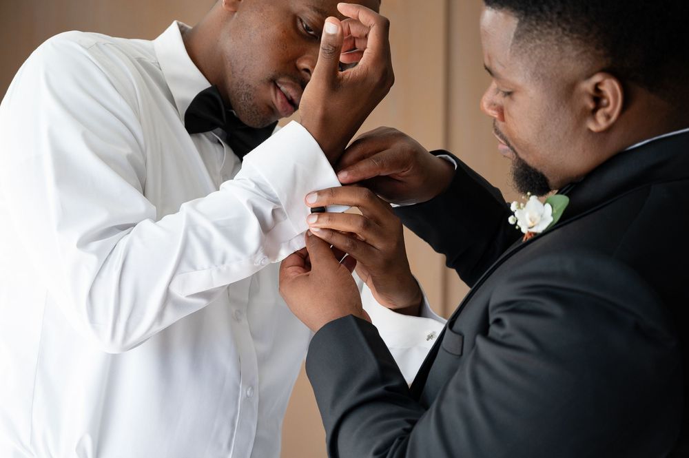 Two people in formal attire help each other get ready for a wedding with one adjusting the other's shirt sleeve.