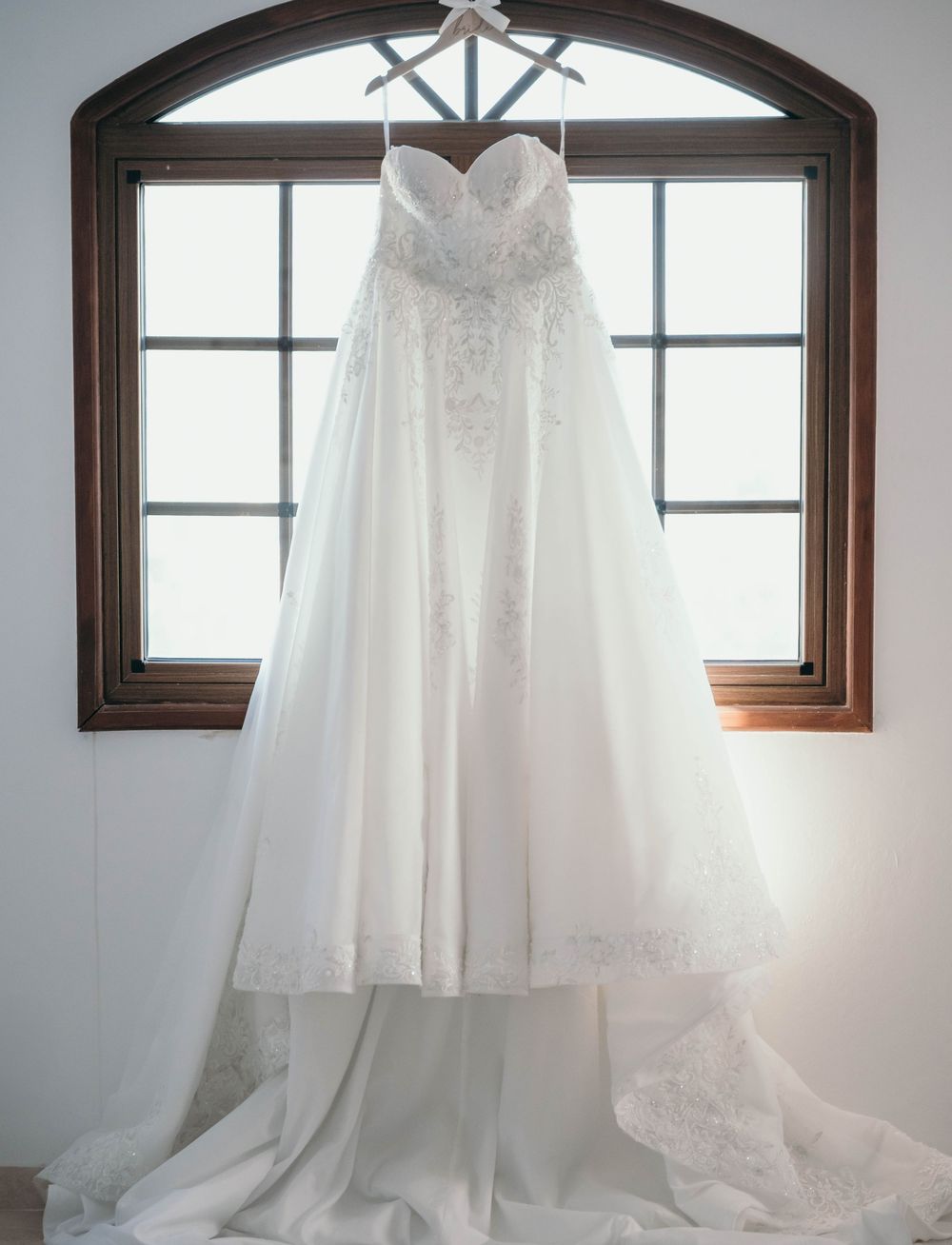 A white wedding dress hanging in front of an arched window with natural light streaming through.
