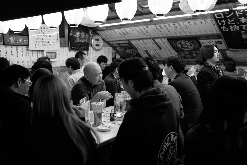 Crowded Japanese izakaya bar with customers seated at counter under dim lighting.