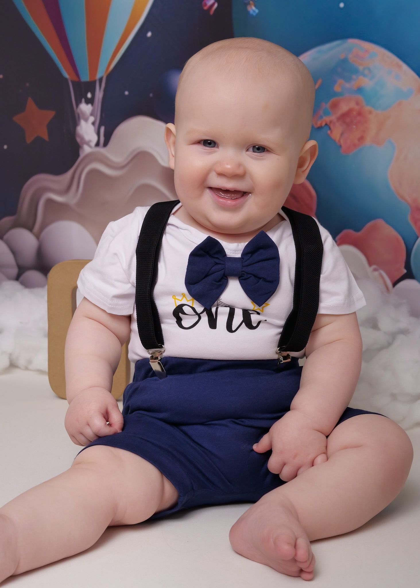 Baby wearing navy blue suspenders and bow tie outfit against hot air balloon backdrop.