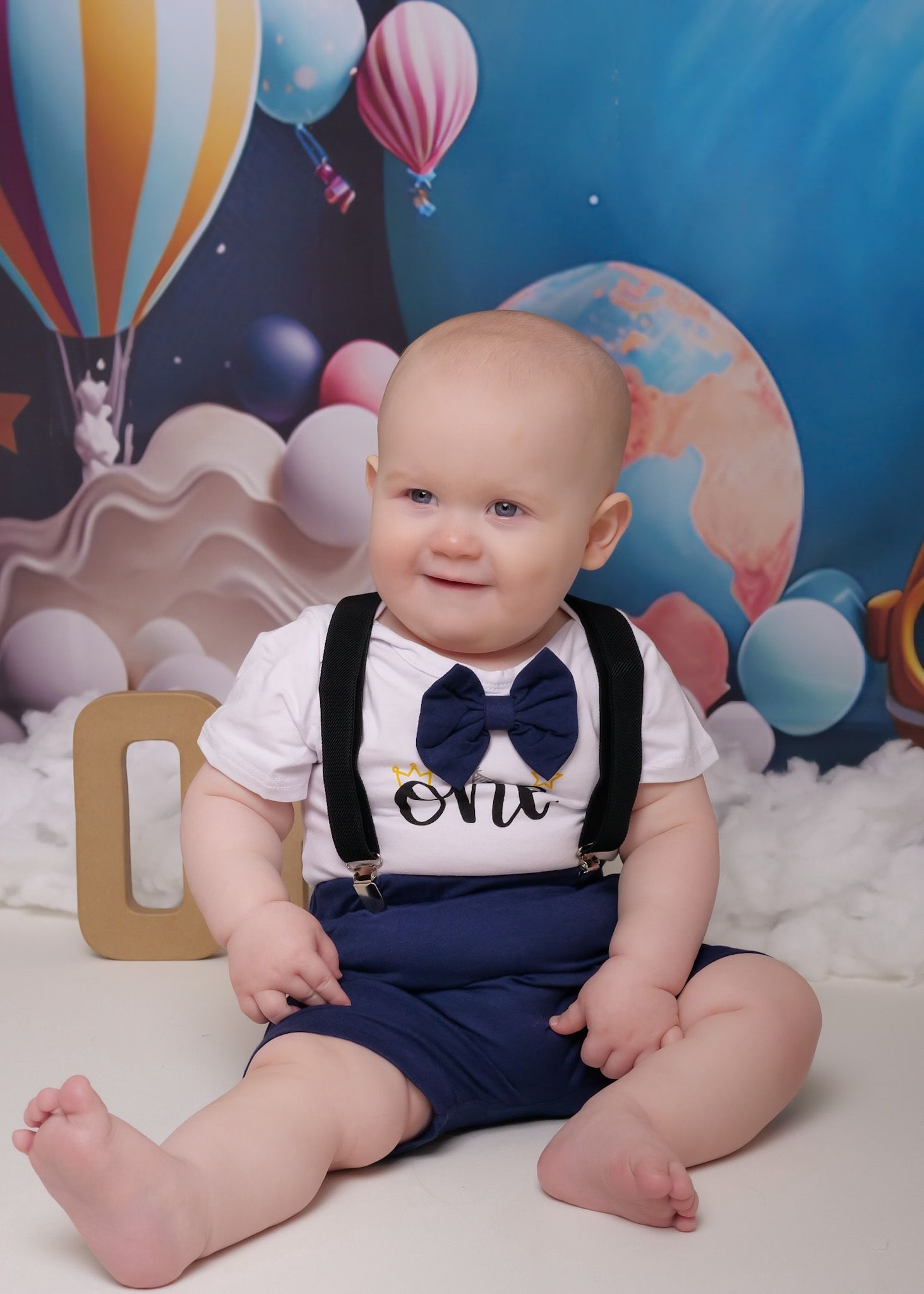 Infant in formal navy and white outfit sitting against colorful balloon-themed backdrop.