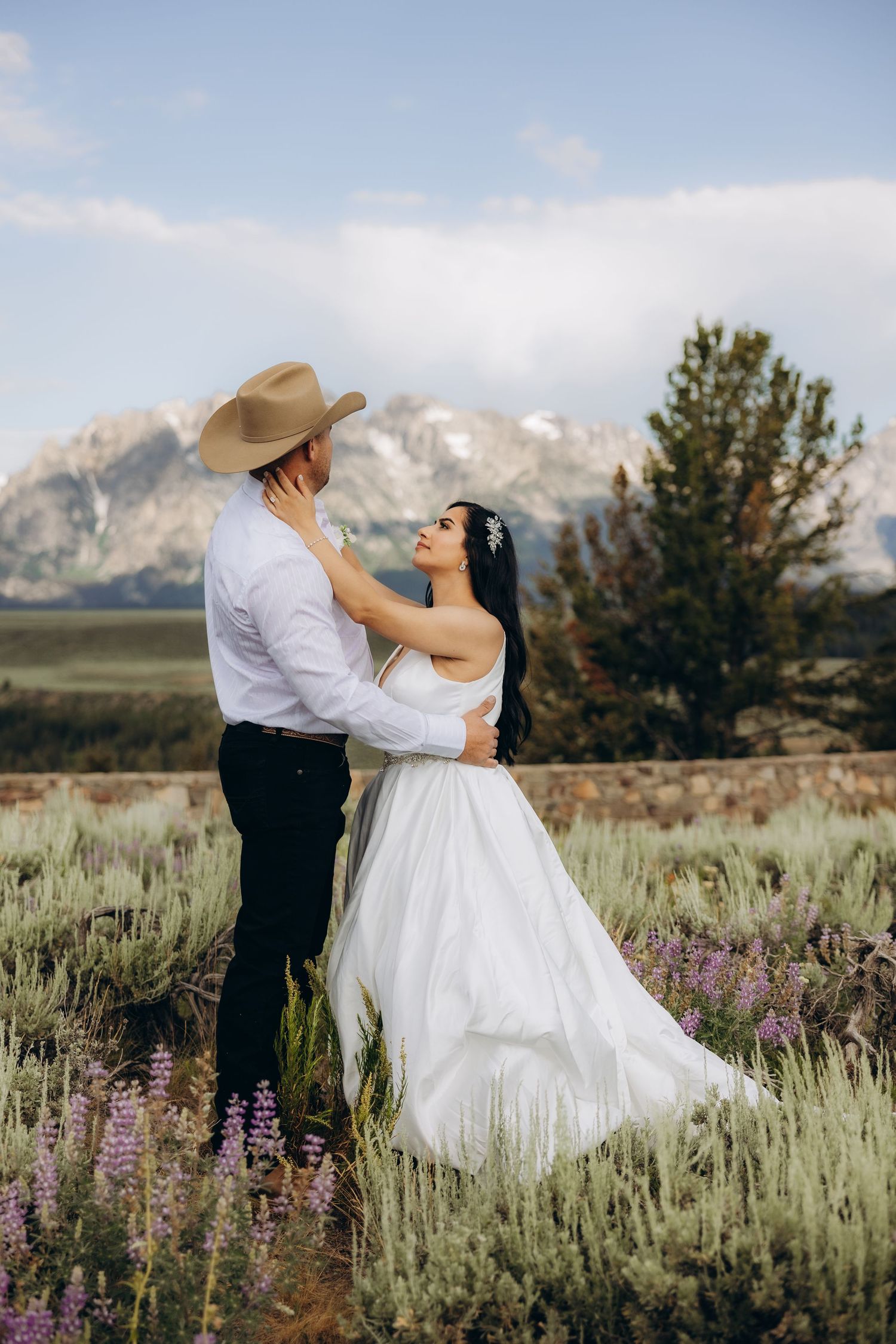A couple in wedding attire embrace in a meadow of wildflowers with majestic mountains in the background.