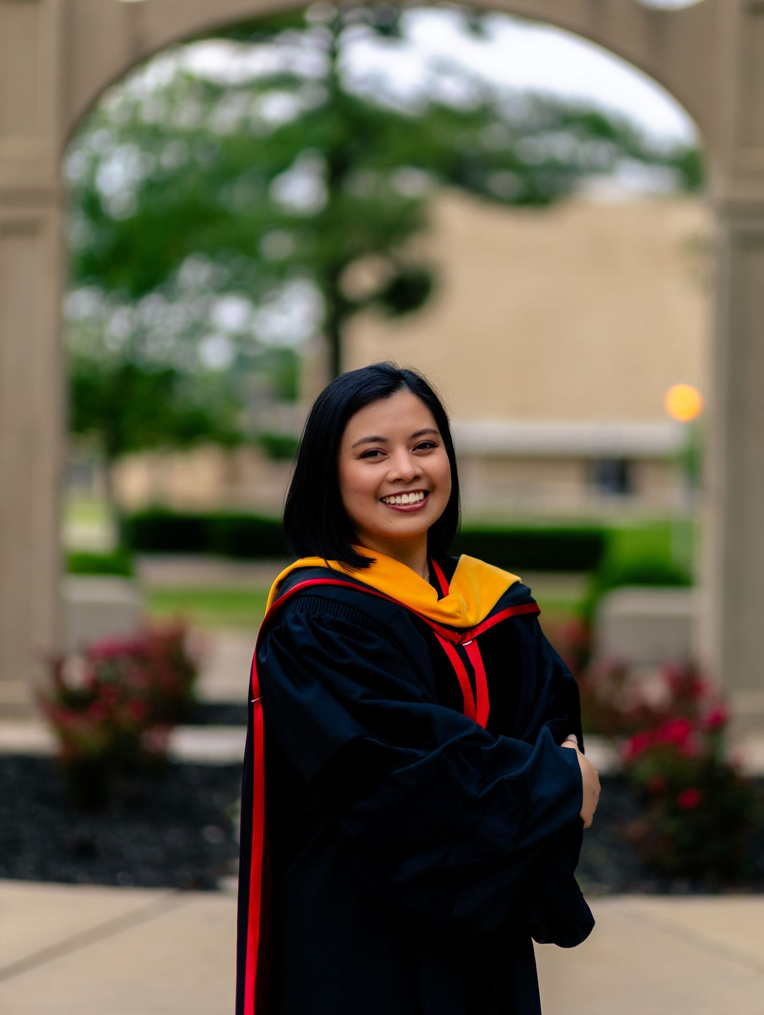 Graduate in black gown stands outdoors with red and gold academic hood against brick building and greenery.