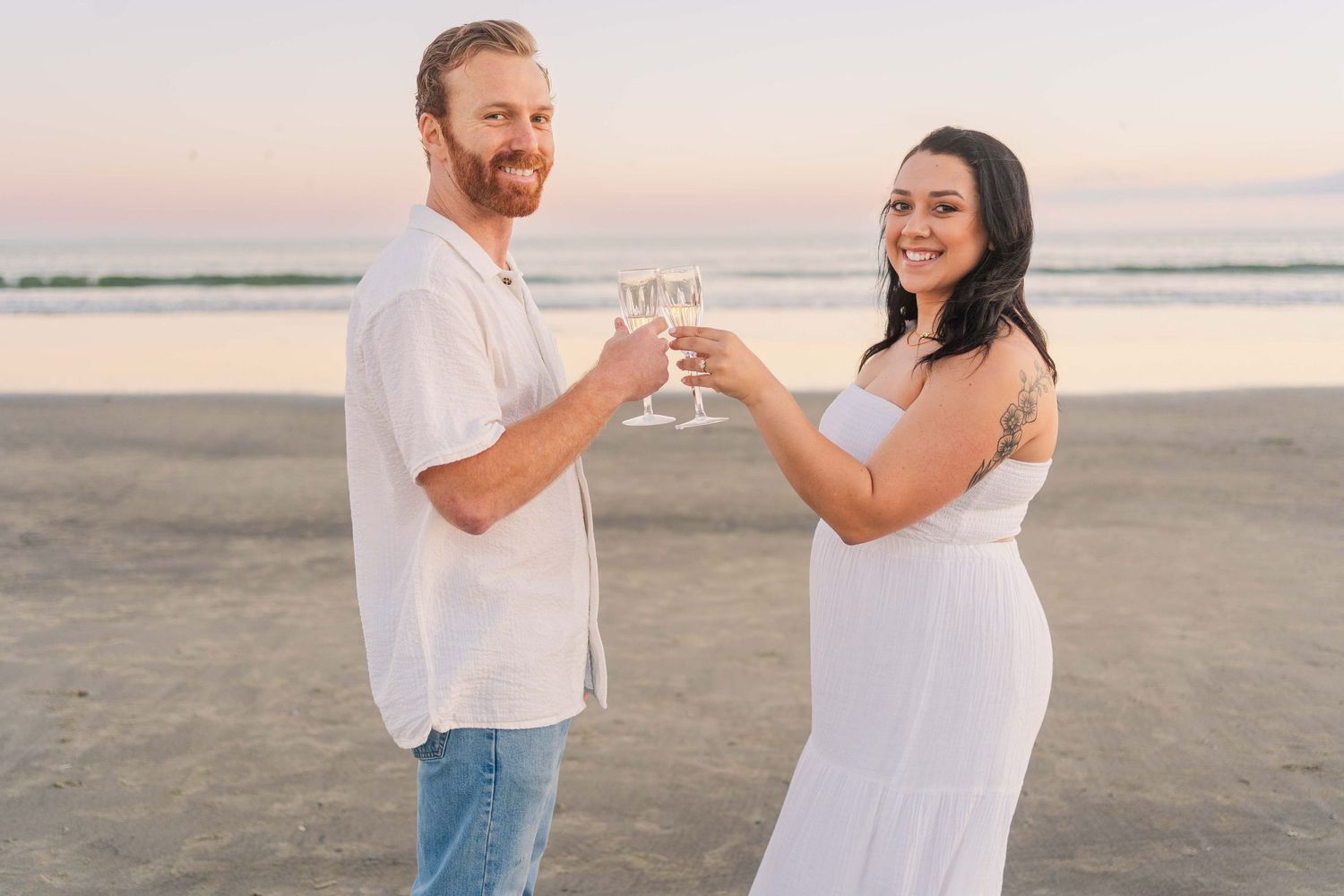 A romantic sunset beach photoshoot showing a couple in casual attire holding champagne glasses at the shoreline.