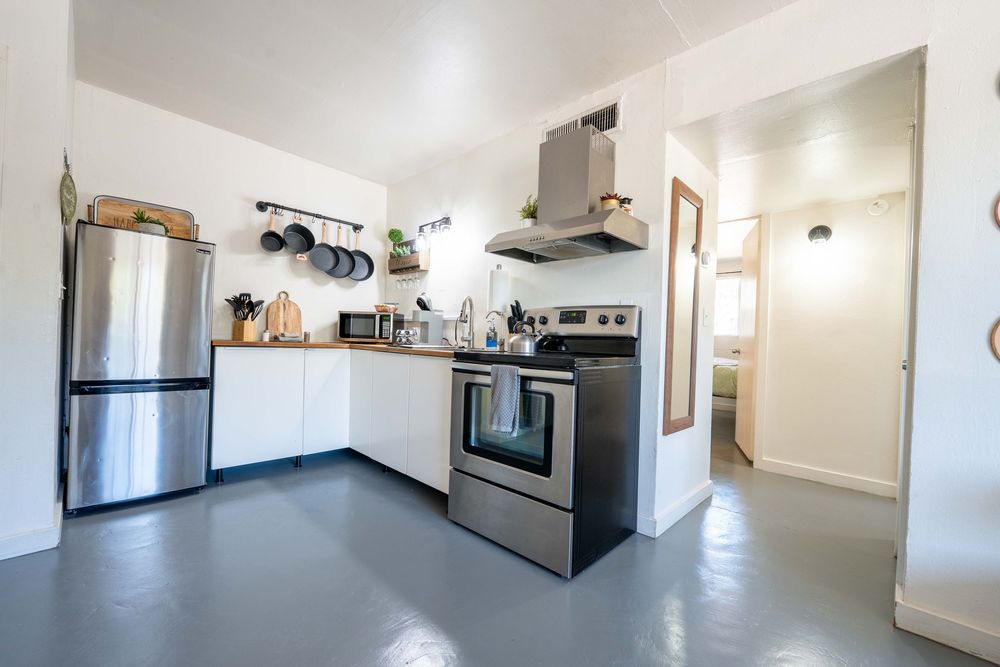 Modern white L-shaped kitchen with stainless steel appliances, open shelving, and grey concrete floors in bright space.