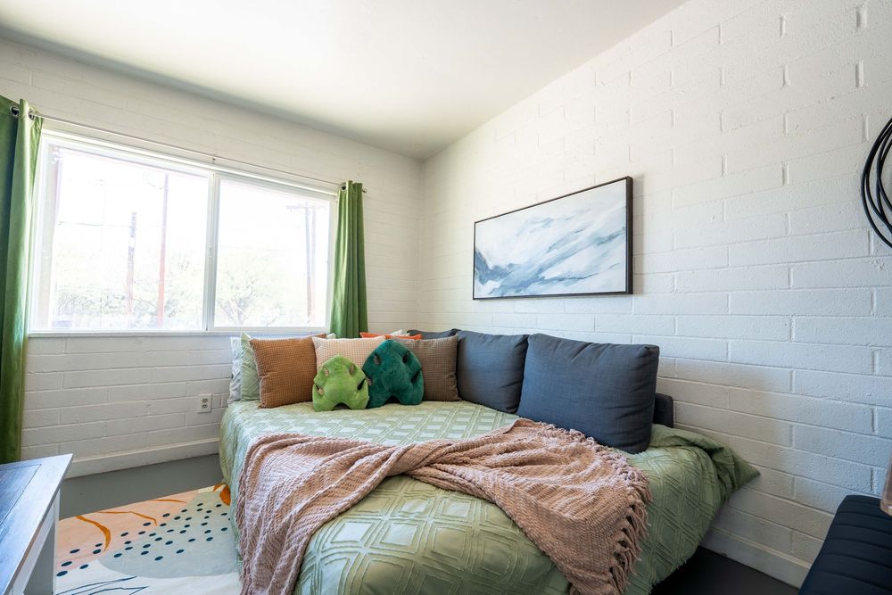Modern bedroom with gray daybed, colorful pillows, green curtains, and framed artwork on white brick walls.