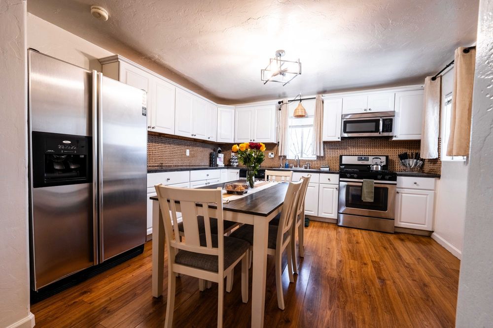 Modern kitchen with white cabinets, stainless steel appliances, hardwood floors and small dining table with chairs.