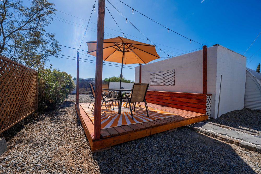 Wooden deck patio with orange umbrella and outdoor furniture on gravel surrounded by white walls and string lights.
