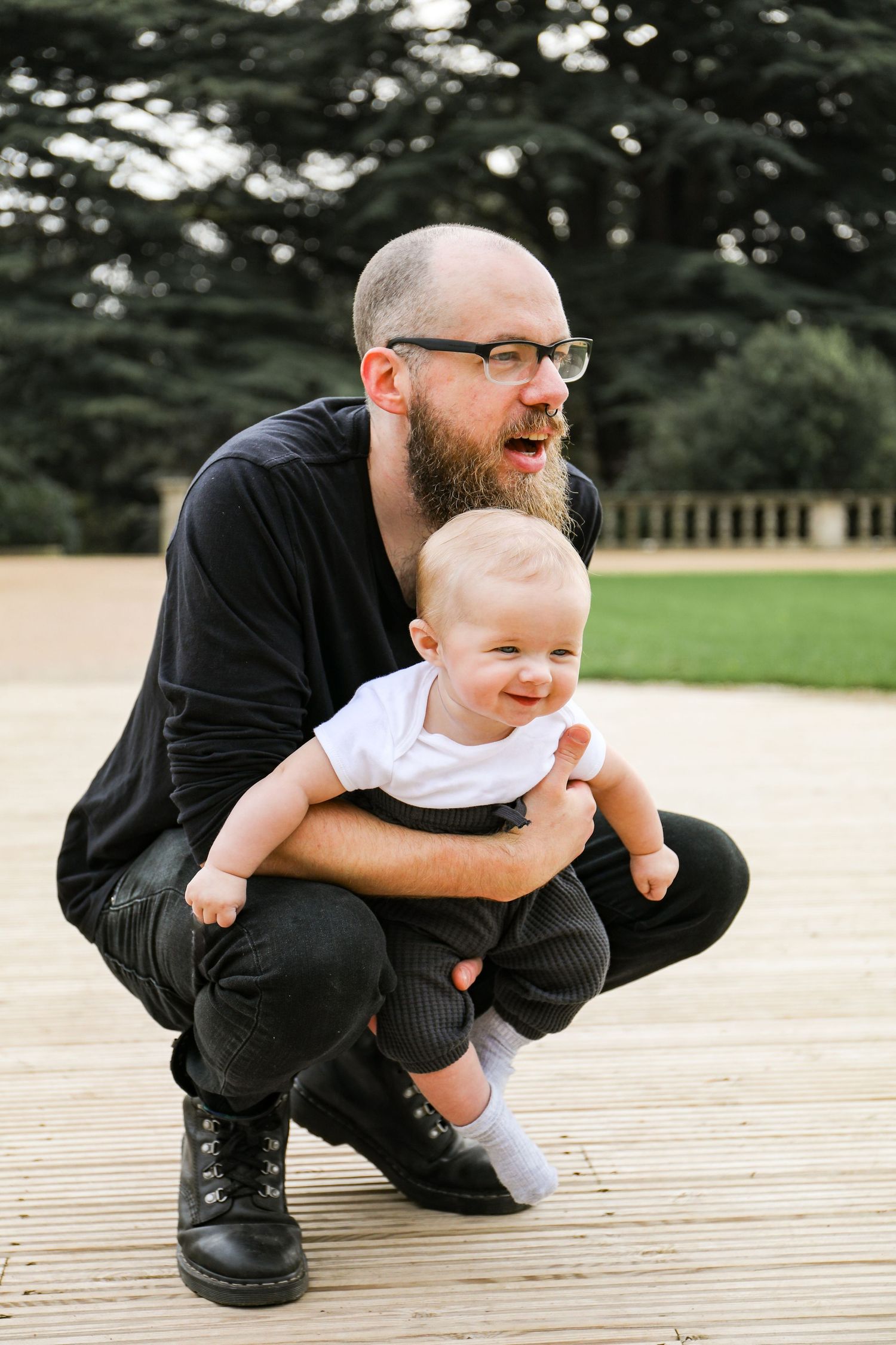 A Dad crouches on a wooden deck outdoors while holding a smiling baby in white clothing, during autumn family shoot in Nottingham