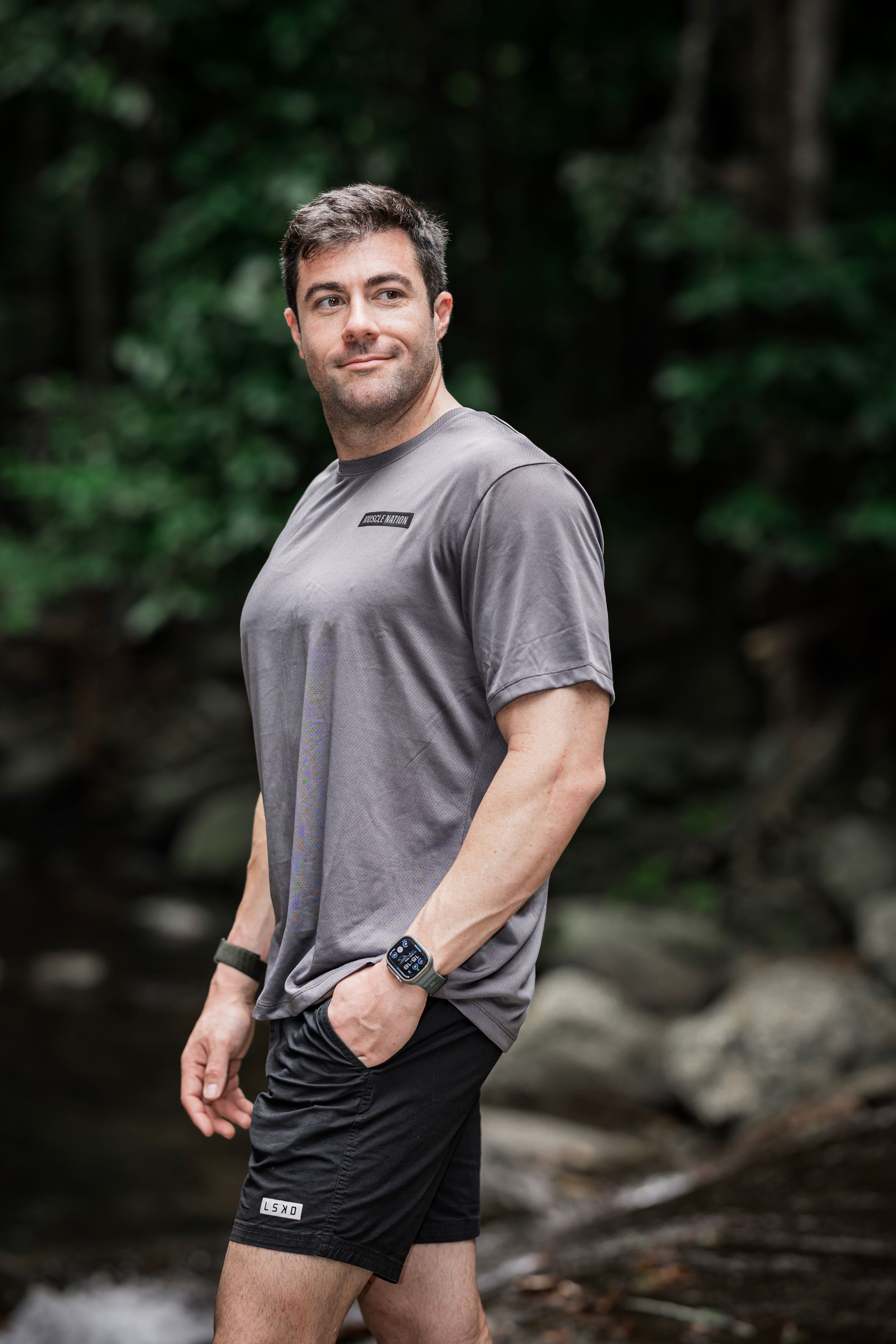 Athletic model wearing grey t-shirt and black shorts poses in outdoor nature setting with wooded background.