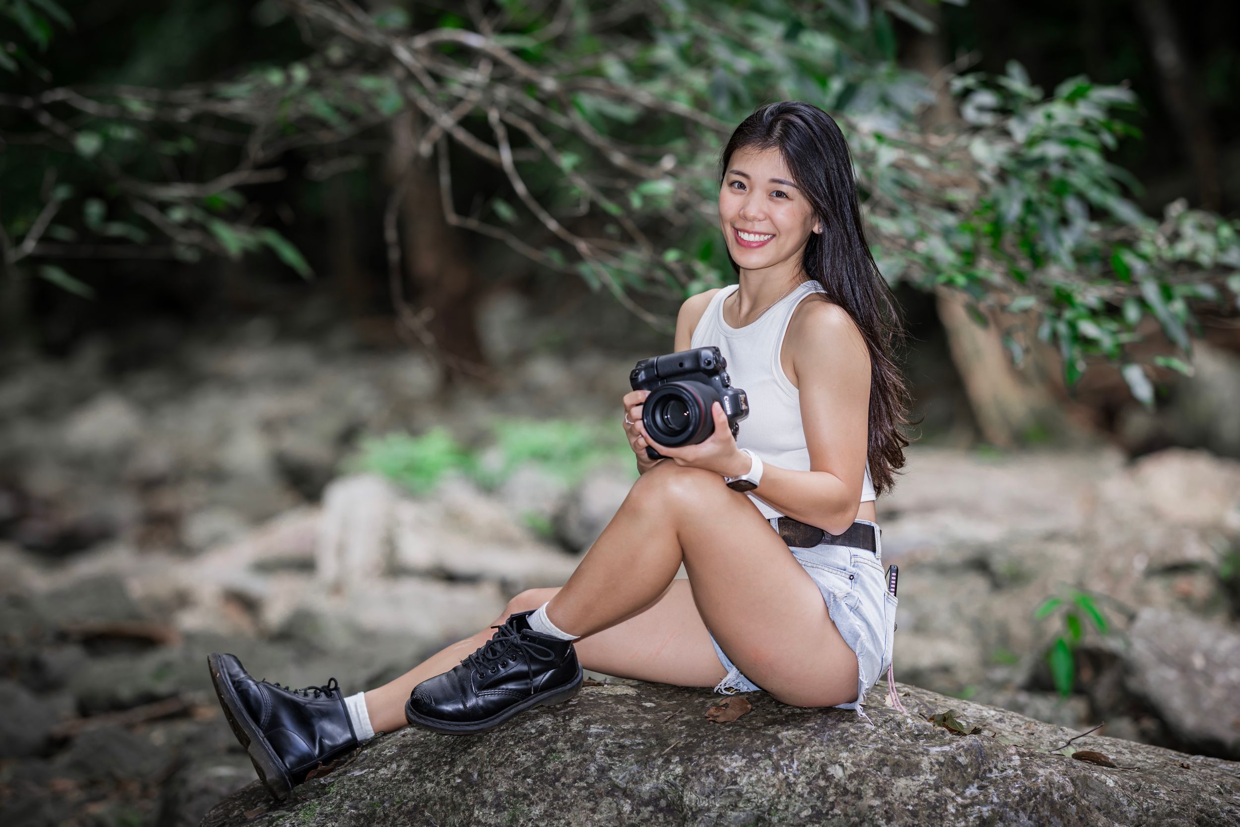 Photographer sitting on rocky ground in nature holding a DSLR camera while wearing casual summer attire.