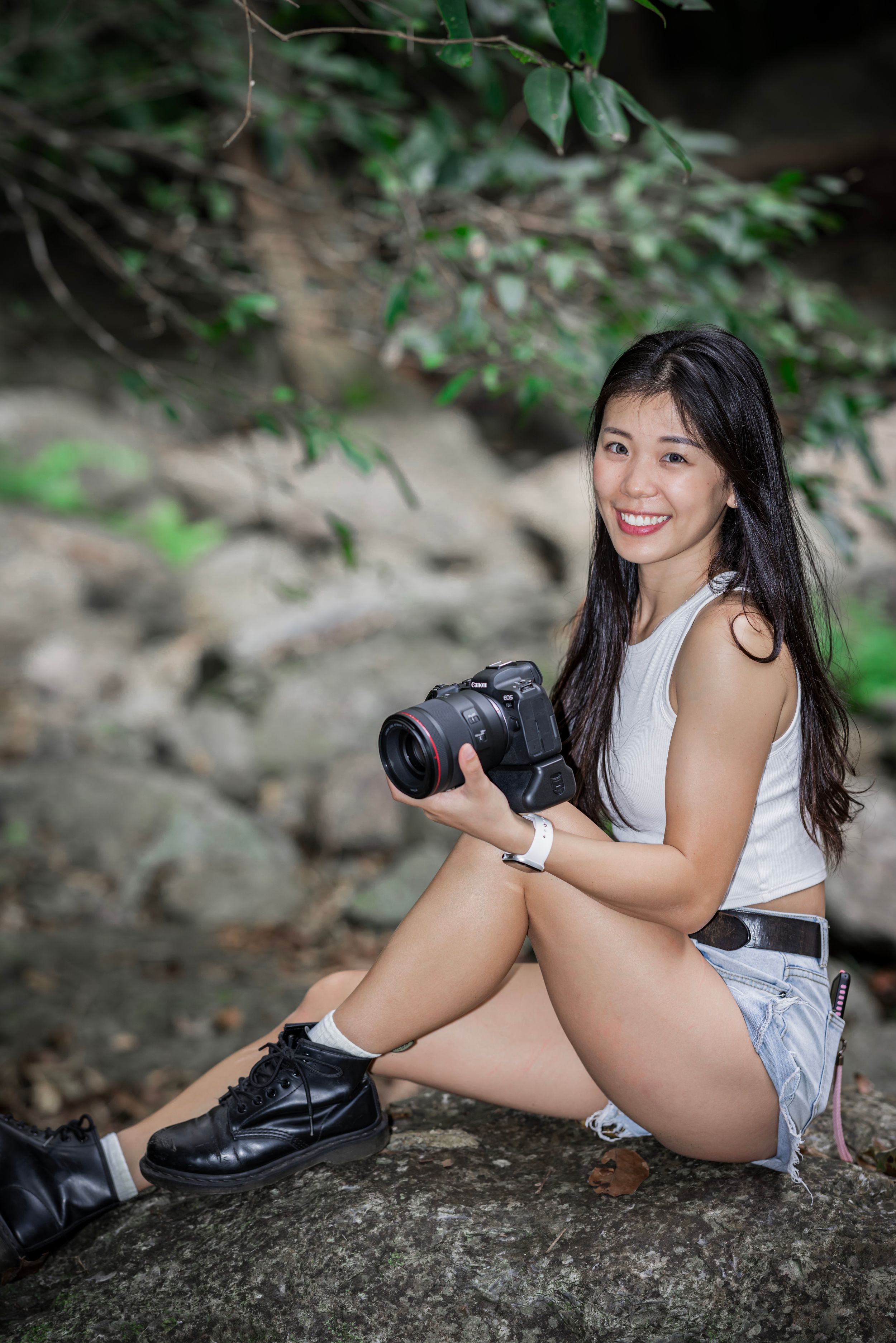 Photographer with camera sits on rock in nature setting wearing white top and shorts with black boots.
