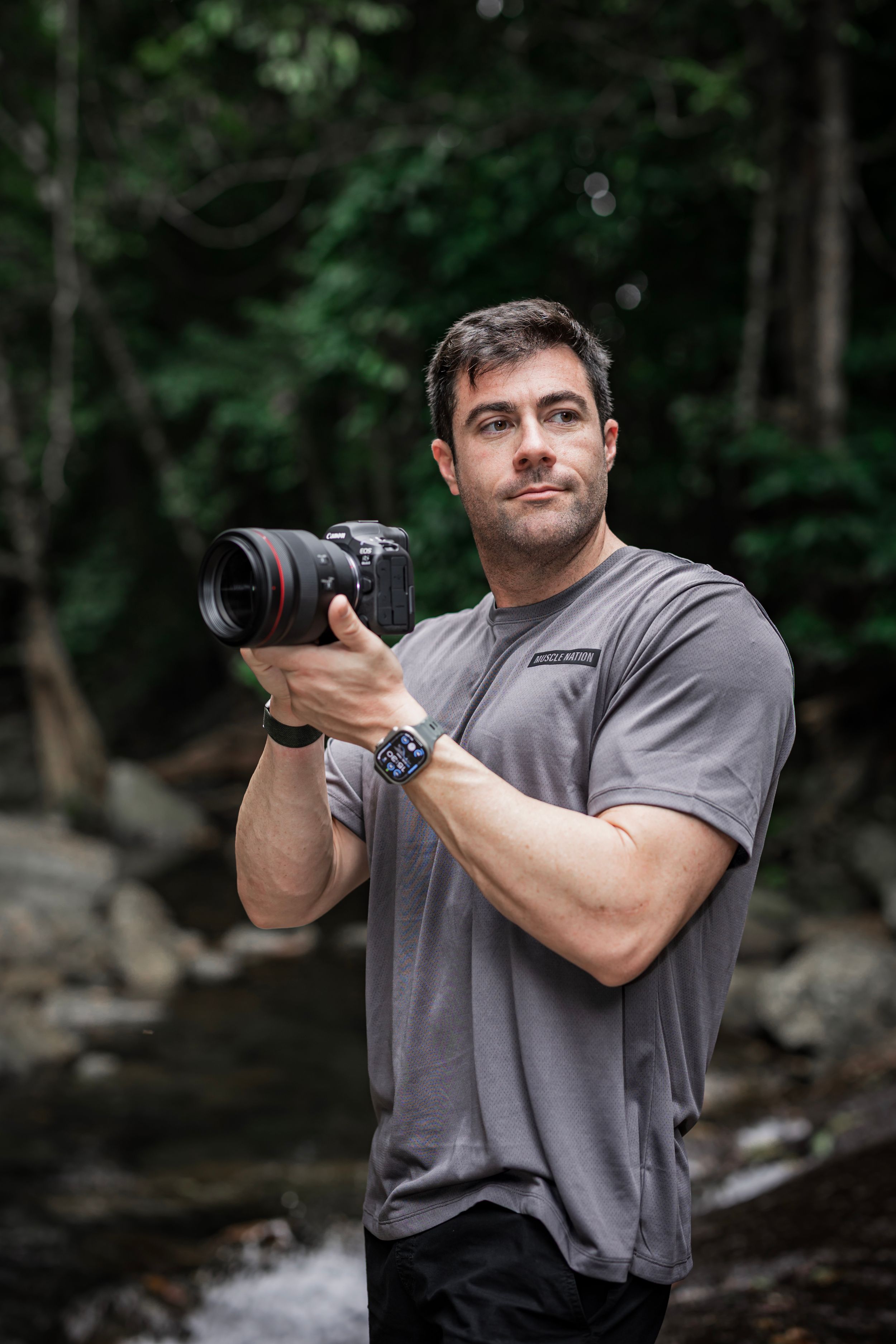 A photographer in a gray t-shirt demonstrates different shooting positions with a camera in a wooded setting.