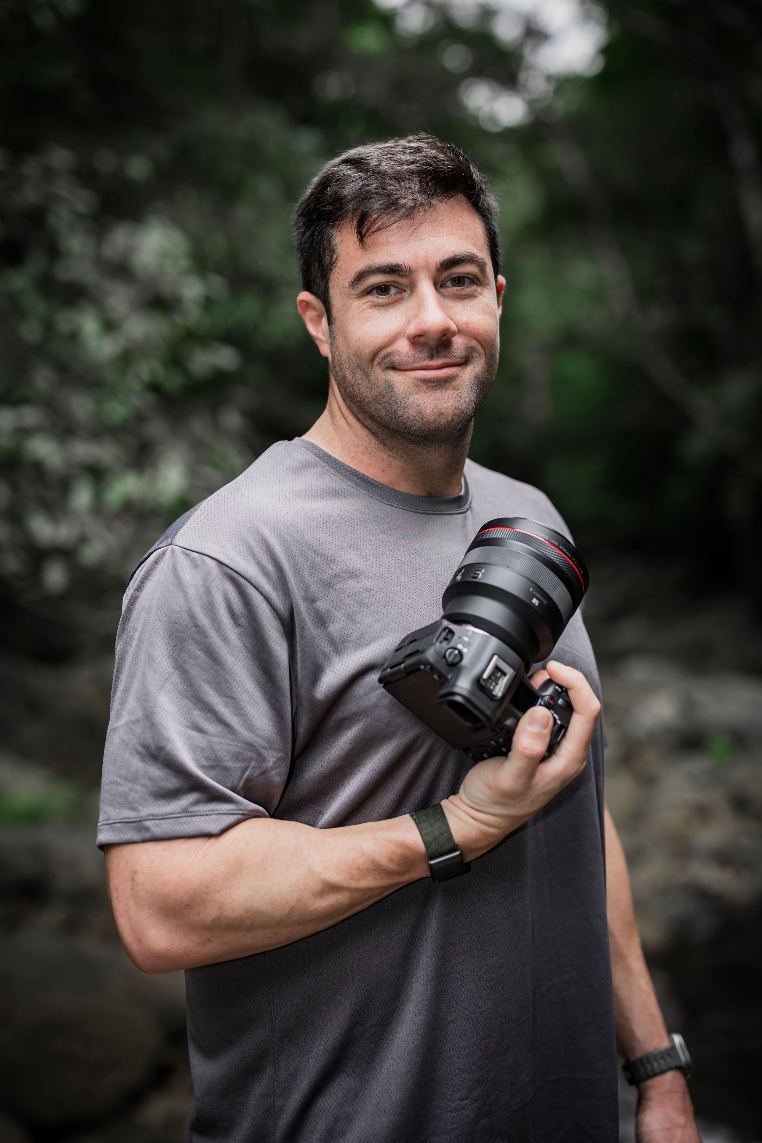 A photographer in a gray t-shirt shows off a professional camera lens while standing outdoors.