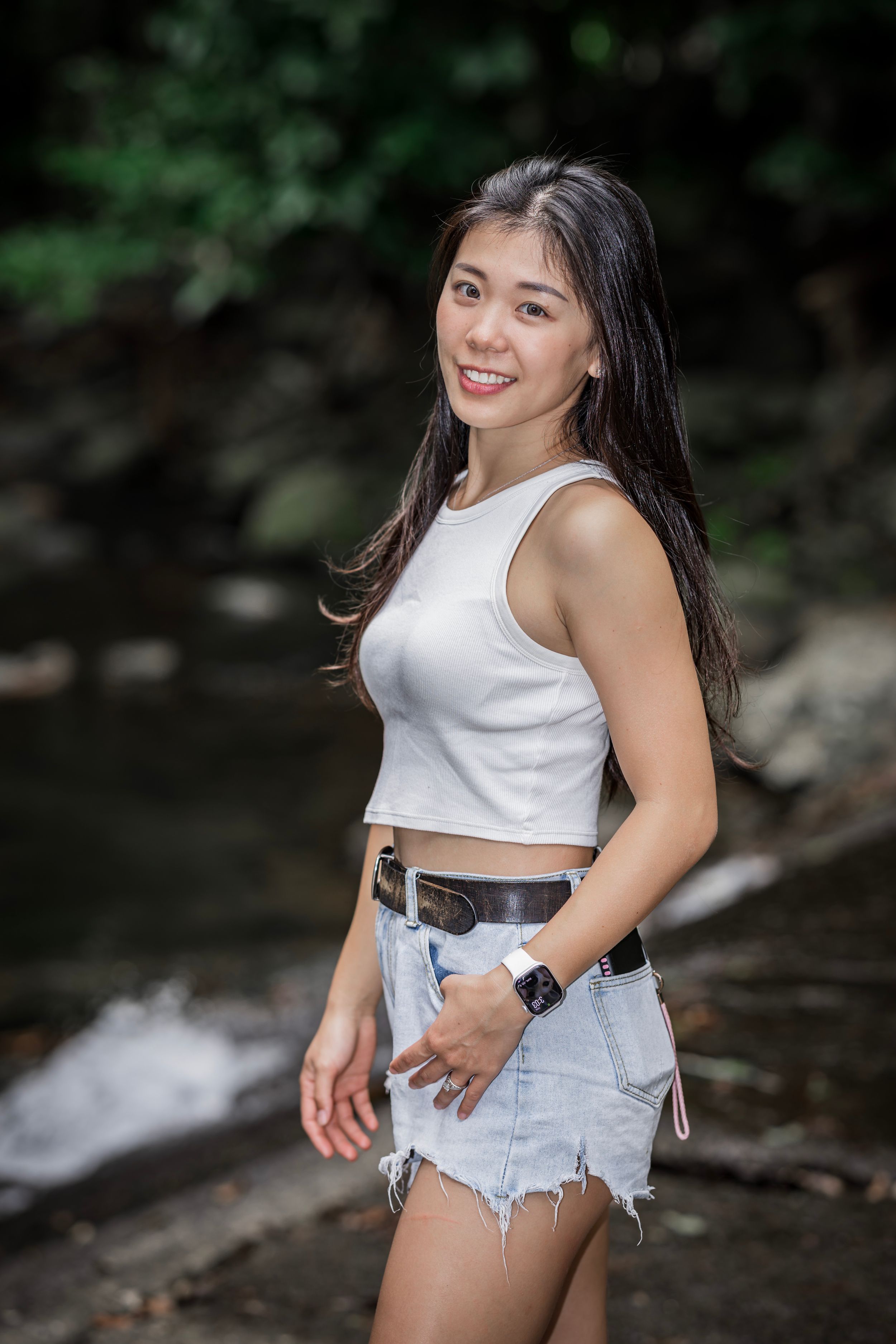 Young woman in white crop top and denim shorts poses playfully in an outdoor nature setting.